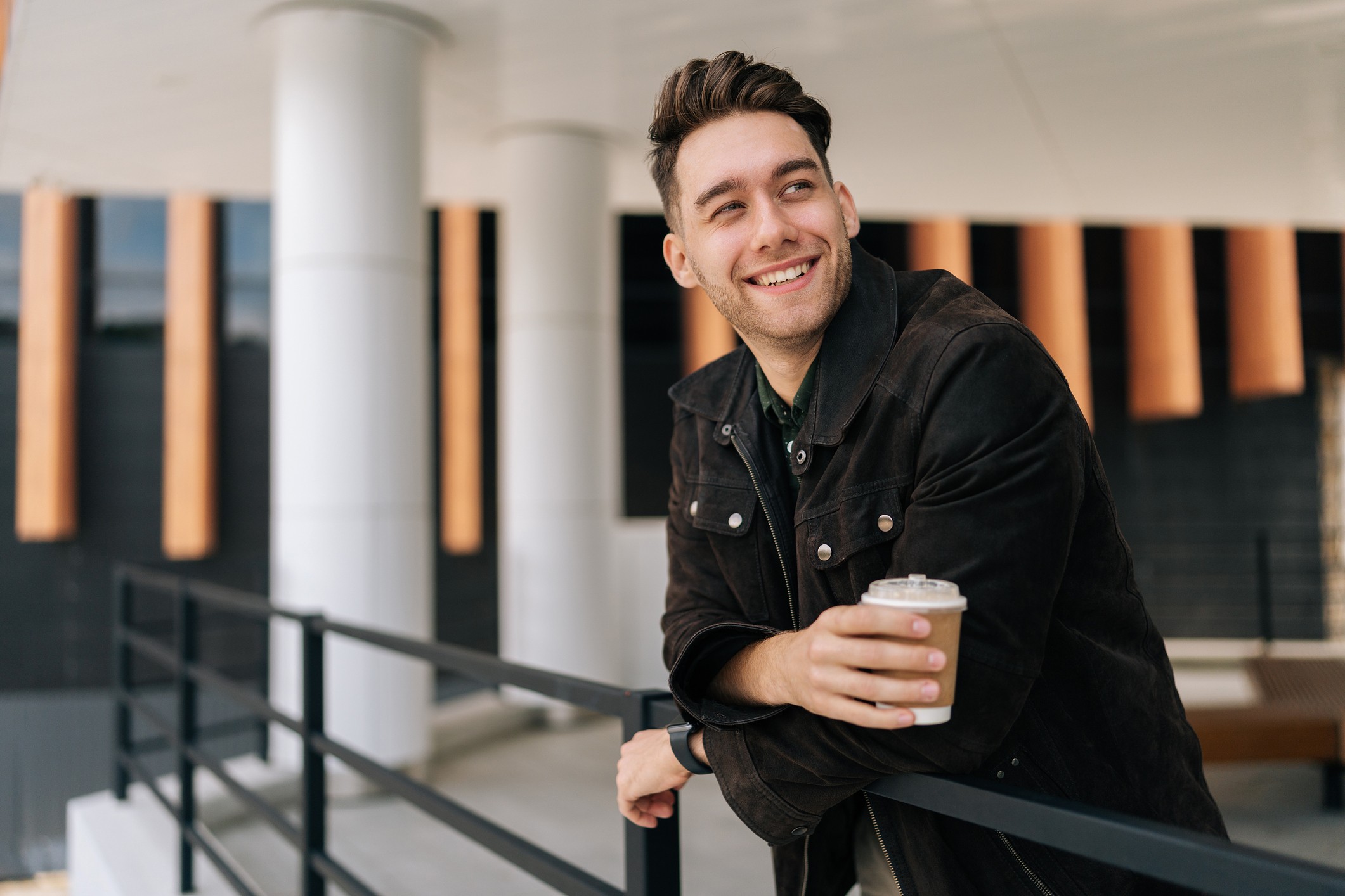 Smiling professional in dark jacket holding a coffee, leaning on a railing in a modern building